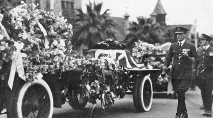 A funeral procession featuring a flower‑covered caisson draped with a Union Jack flag. Uniformed servicemen walk alongside the carriage as it moves along a street lined with palm trees and large buildings with towers in the background.