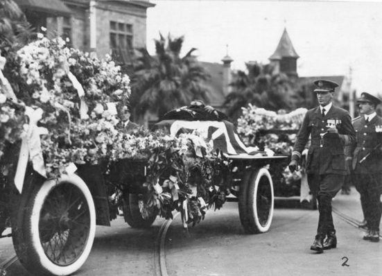 A funeral procession featuring a flower‑covered caisson draped with a Union Jack flag. Uniformed servicemen walk alongside the carriage as it moves along a street lined with palm trees and large buildings with towers in the background.