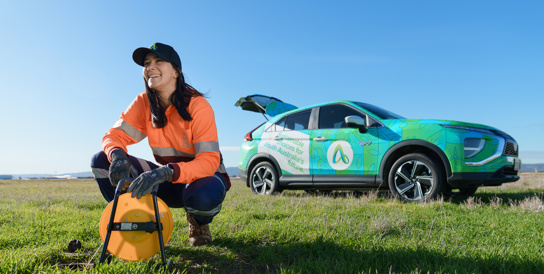 woman in hi vis crouches on grassed are infront of branded ev car