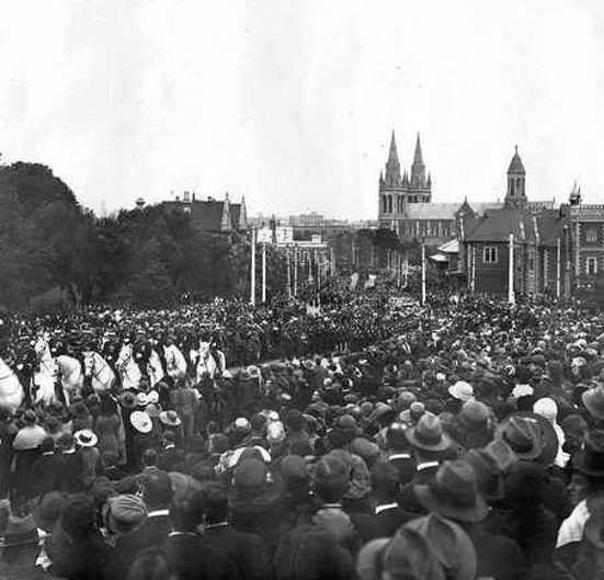 A large crowd fills a public square, watching a ceremonial procession led by a team of white horses pulling a carriage. People wearing hats stand closely packed in the foreground, while the procession and uniformed participants move through the center. In the background are tall church spires and historic stone buildings, creating a prominent skyline.